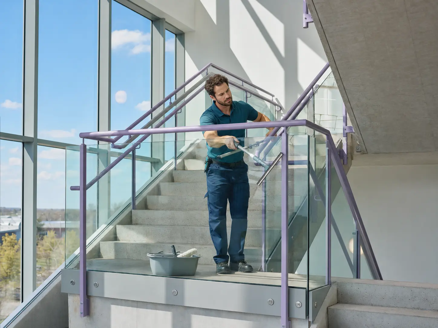 An operative squeegeeing a glass balustrade on an internal stair landing beside a full-height window.